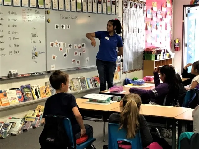 young children sit in desks while a teacher in a blue shirt stands at the white board where different food items are displayed