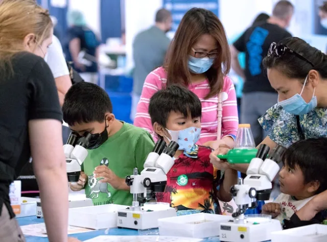 Three dark-haired kids assisted by two women use microscopes.