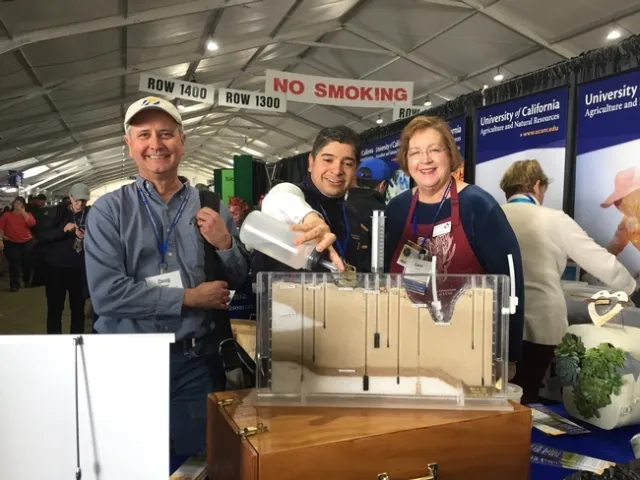 Man standing between a man and woman pours water into clear plastic demonstration kit filled with sand