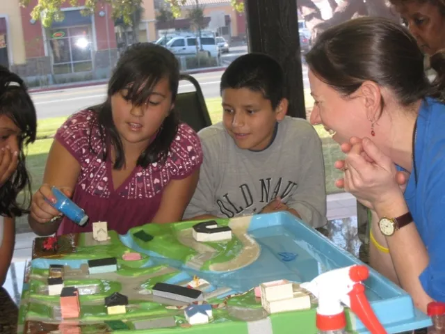 Two students watch as a girl squirts water onto a plastic model community. Drill, on right, smiles as she observes.