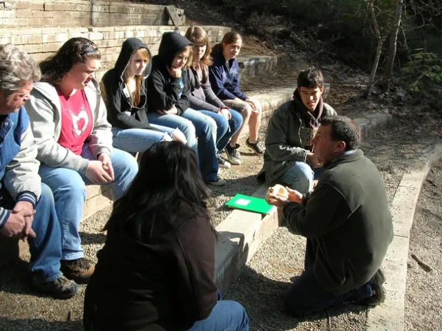 Borba kneels down to face a group of 8 young people while holding what appears to be an animal bone.