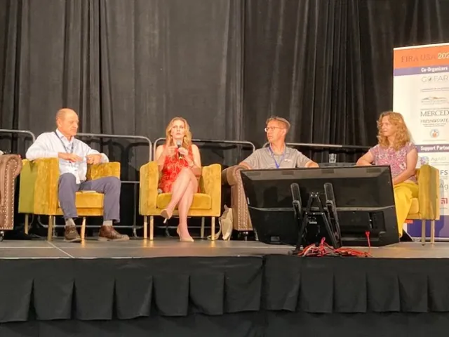 Four people sit in armchairs on a stage. Bettencourt speaks into a handheld mic.