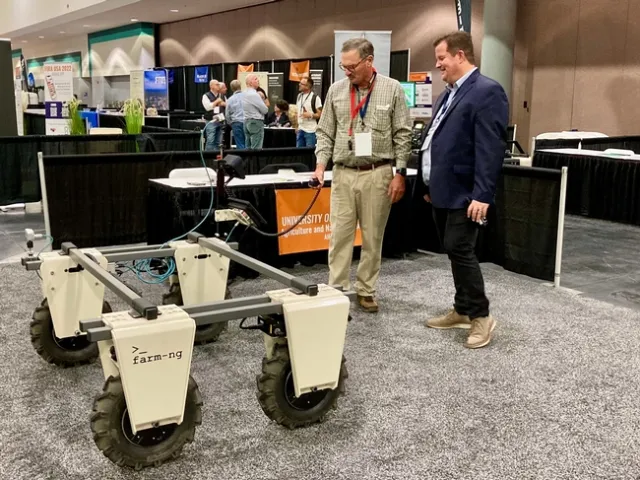 Fennimore controls an automated platform with a handheld device connected to the machine as Youtsey observes. Equipment vendor booths are in the background.