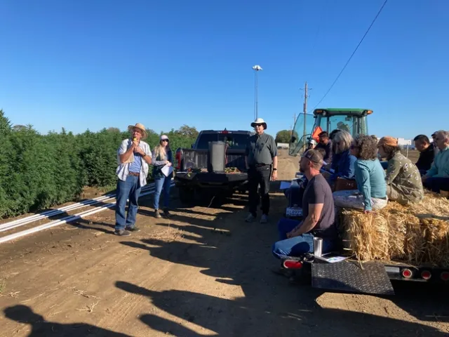 Standing beside a field of hemp, Bob Hutmacher speaks into a mic as growers and others sit on hay bales listening.