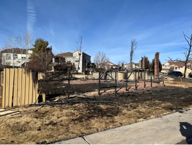 Fire scorched grass black in the foreground. A few yellow boards of the fence remain standing beside the burned posts.