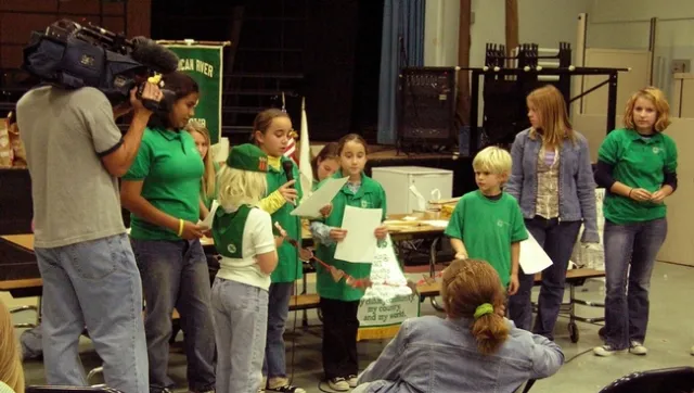Kids in green shirts read from pieces of paper as someone shoots video.