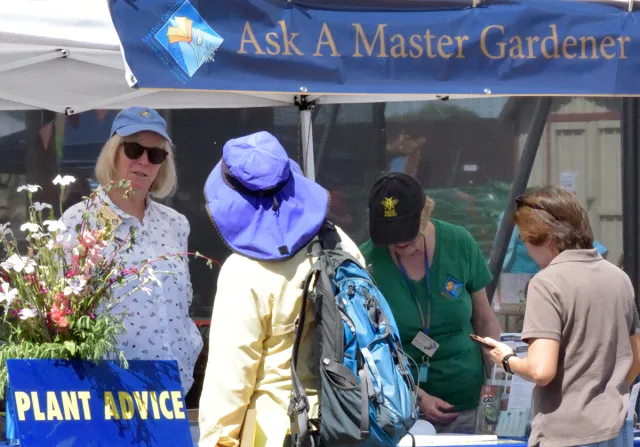 A booth labeled "Ask A Master Gardener" features three women discussing plant advice. One is wearing sunglasses, another a green shirt and black cap, and a third wears a blue hat and yellow jacket with a blue backpack.
