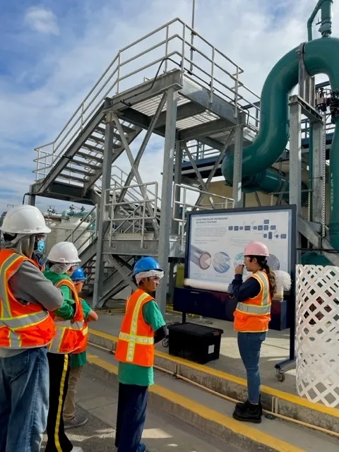 A young woman stands in front of a poster, presenting information to her audience of young students.