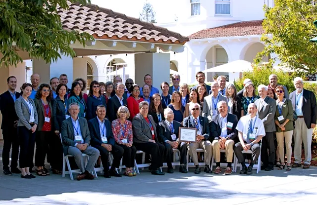 A group of people sitting in a row of chairs and two rows standing behind the chairs. Soroosh Sorooshian holds plaque next to Richard Rosenberg.
