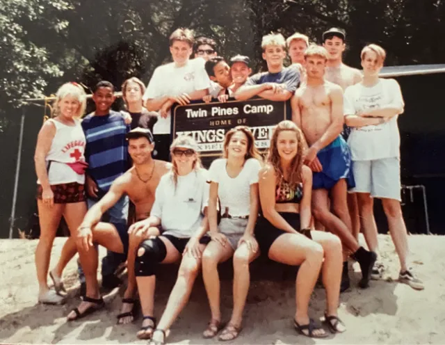 A woman poses on the beach with a group of teenagers in front of a beach volleyball court.