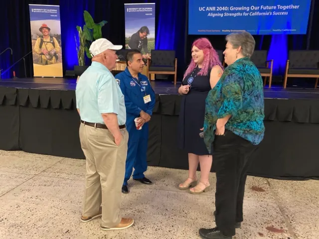 Four people stand in a circle conversing with UC ANR banners and stage in background.
