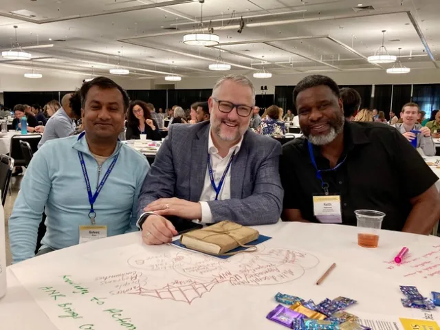 Three smiling men sit at a round table in a conference hall.