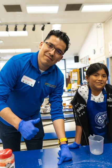 One adult and one youth in a school cafeteria with food prep gloves on