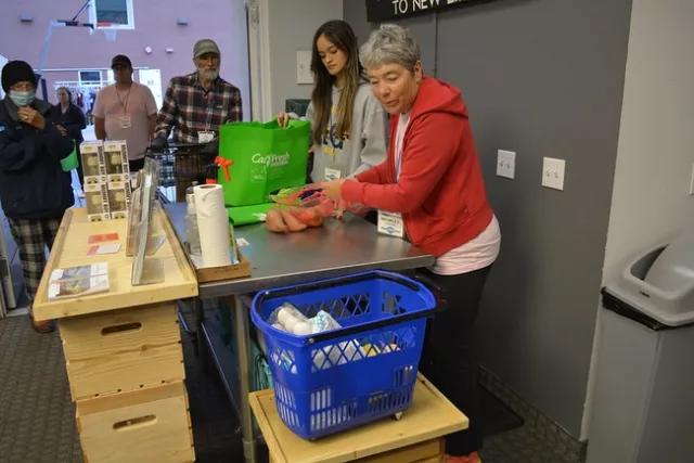Two volunteers at a counter handling groceries while customers wait in line for food