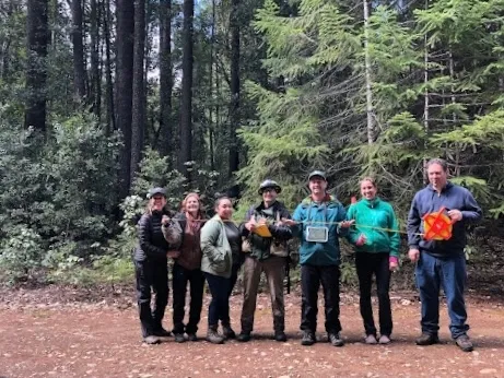 A group of seven people standing in a row in a forest clearing
