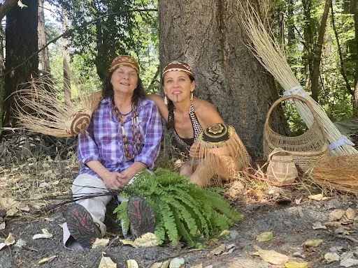 Two women sit at the base of a large tree trunk surrounded by basket and basket-making materials.