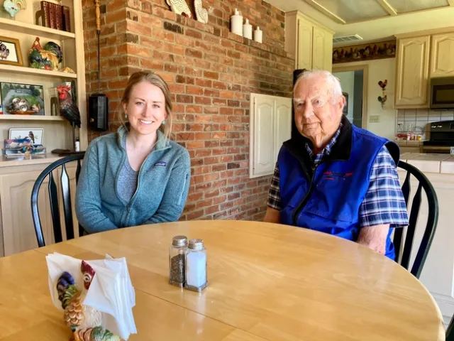 Grace and Jack sit at a kitchen table with a ceramic rooster napkin holder on it. Rooster decorations hang on the walls and sit on shelves behind them.