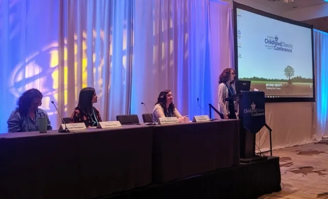 Gail Woodward-Lopez speaks at a podium at a childhood obesity conference. On left, three panelists sit at a table.