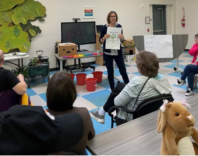 A classroom with students and a volunteer holding a sign that reads “Parts of a Plant.”
