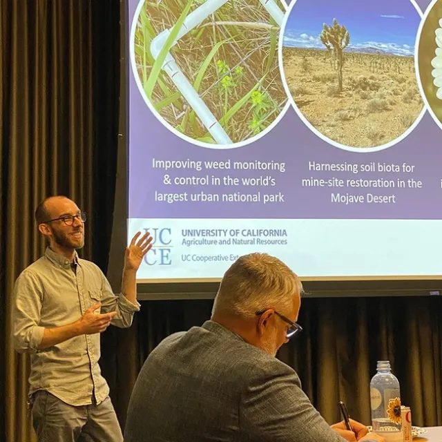 Justin smiles as he gestures toward a slide with images of weeds. 