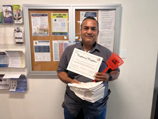 A man, holding papers and a fly swatter, smiling at the camera.
