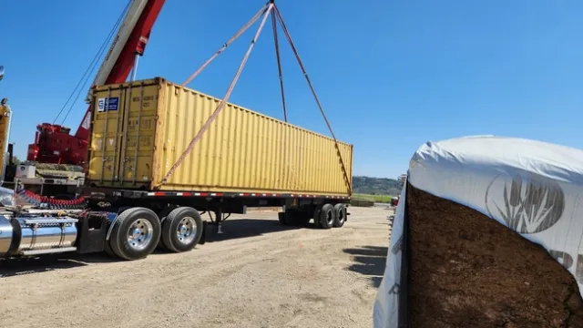 A crane loads a yellow shipping container filled with farm equipment onto a flatbed trailer