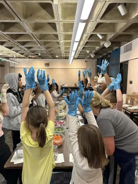 A table set-up with food and youth with gloves on and hands in the air. Adults providing instructions