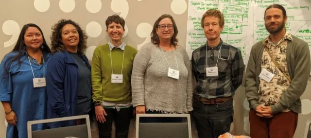 Six people wearing name tags stand in front of a wall that is partly covered in white poster paper with discussion notes written in green ink.