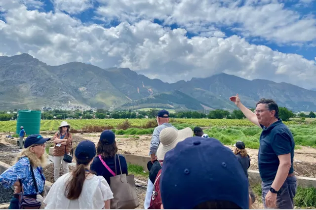 A man gestures toward a green crop field as tour participants listen to him speak.