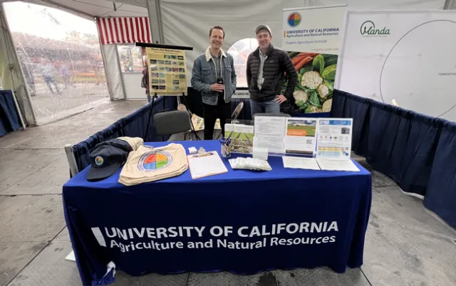 The two men are standing behind a table of OAI logo hats and bags and brochures.