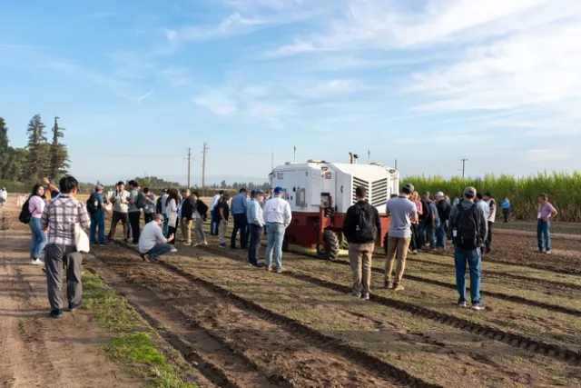 Dozens of people gather around a large machine rolling through an unplanted field and