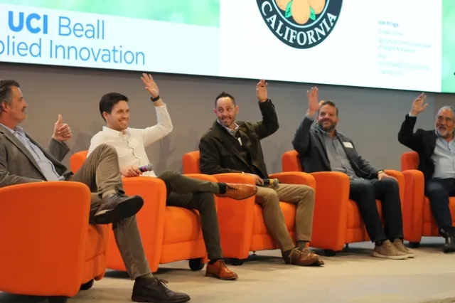Five middle aged men sit on stage, raising their hands.