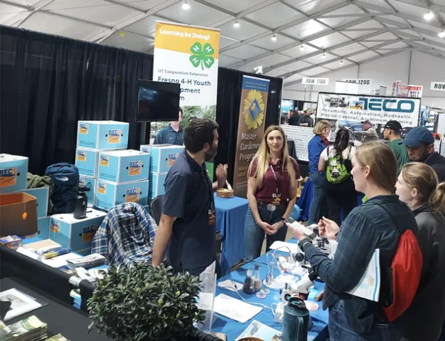 Brady and Lauren stand behind an exhibit table with microscopes while talking with two people.