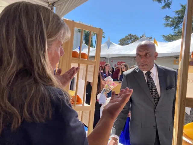Tammy stands behind the booth table and Bradford picks up an avocado