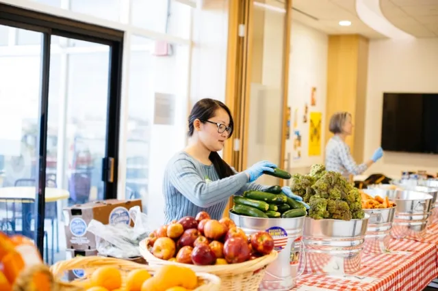 A woman wearing blue gloves sacks zucchini in a metal bucket on a table of baskets of oranges, apples, broccoli and carrots.