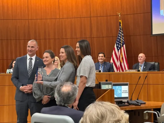 Four people pose for a photo with Shannon holding up the lucite trophy as the board of supervisors look on in the background.