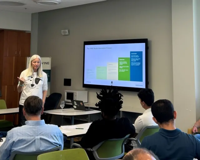 A woman speaks to a group of people sitting at desks. The slide on the screen reads: The VINE Studio Innovation Process.