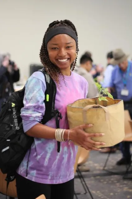 A young African American lady holds her tomato plant while smiling big.