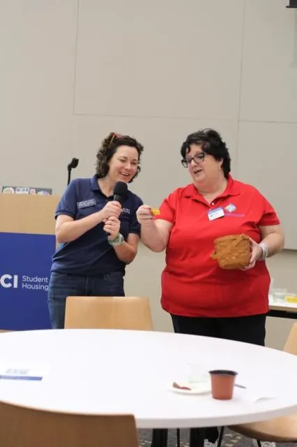 Two middle-aged women. One holds a basket with raffle tickets and the other has a mic in her hand.