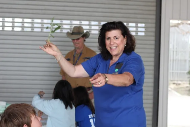 A middle-aged woman holds a chive in one hand and points to a student with the other.
