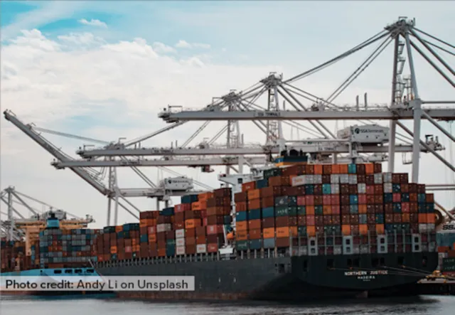A ship loaded with shipping containers sits in port beneath cranes.