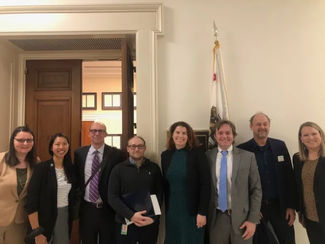 Eight people stand outside of a congressional office. Open door and flag in background.