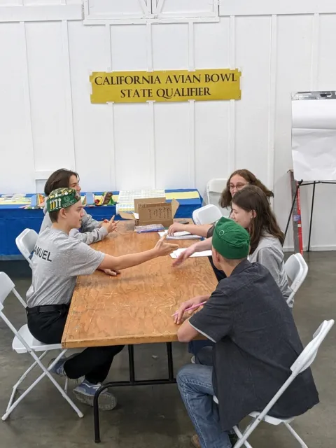 5 youth sit at a table with a sign on the back wall reading, 