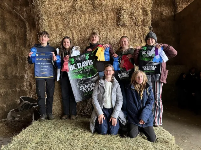5 youth holding ribbons and posters stand in front of hay bale backdrop. 2 youth kneel in front of them.