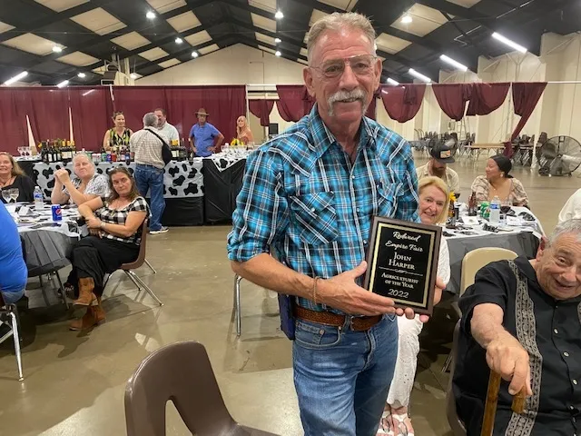 Harper, wearing a blue plaid shirt, holds a plaque that reads, 