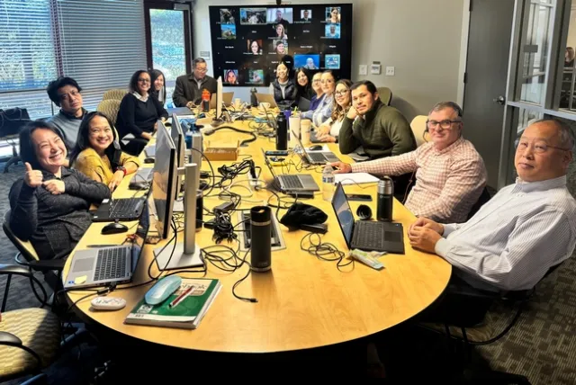 In a conference room, 14 people sit around a conference table littered with laptops.