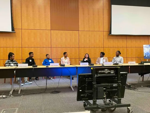 Six youth and Eve sitting in a row against a wall, with tables in front of them. Large camera in foreground.
