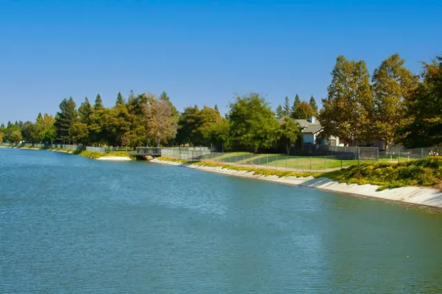 A canal of water next to a fenced area with residential homes and trees.