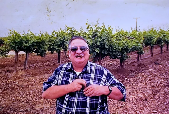 Luvisi, wearing a plaid shirt, stands, grinning, in a vineyard.