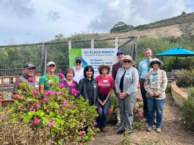 Ten people stand next to a flowering hot pink geranium bush. Hanging on the fence behind them is a sign that reads,